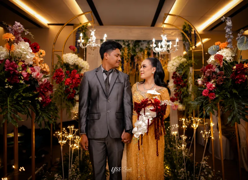 Pasangan berpegangan tangan di antara bunga dan lengkungan emas